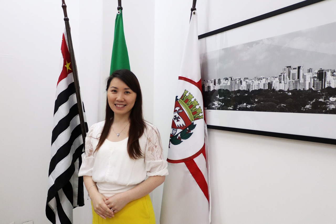 A representante do Conselho de Imigrantes, Yao Na Kim, posa para a foto com a bandeira de São Paulo de fundo